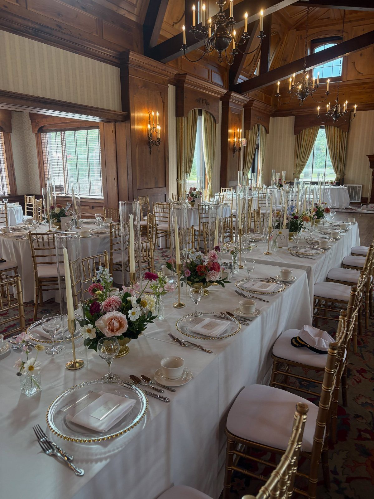 Long wedding reception table decorated with white linens, gold chairs, and small wildflower floral arrangements.