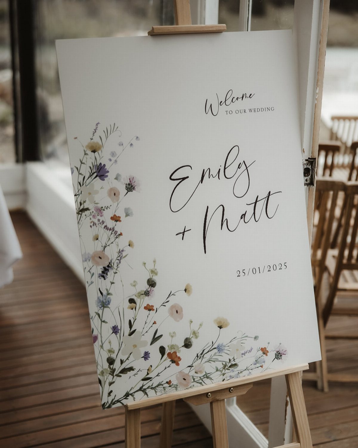 A white wedding welcome sign with wildflower illustrations and names on a wooden easel.